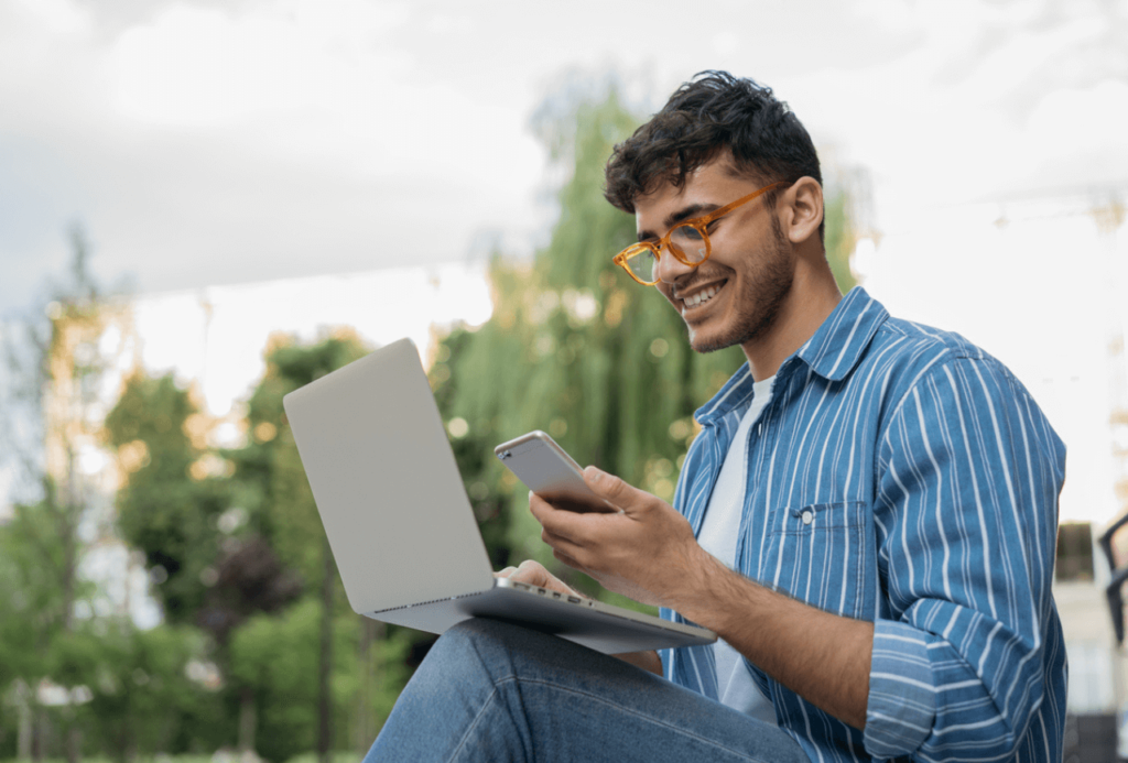homem sorrindo segurando um celular e um notebook. Ele está estudando no exterior à distância.