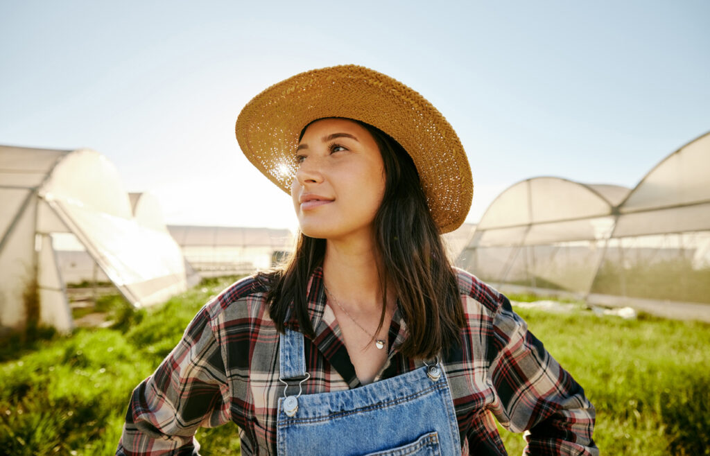 Mulher de chapéu de palha usa uma camisa xadrez e um macacão, olhando para o lado e com um leve sorriso. Ao fundo, há uma plantação com aparentes estufas.