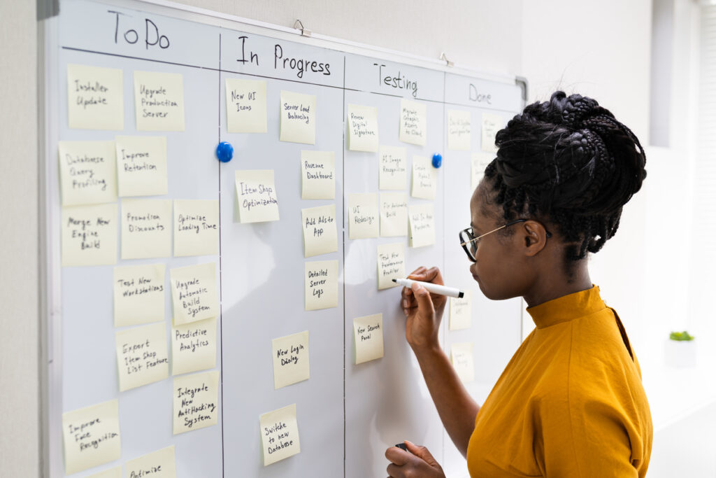 Mulher negra anota em post it em uma parede cheia de post its organizados segundo o kanban.