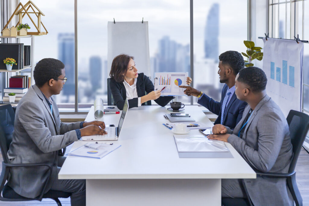 4 pessoas se reúnem em mesa de reunião e uma mulher segura um gráfico apontando informações
